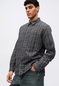 Siva karirana srajca z gumbom, ki ima ovratnik, dolge rokave in mehak material. Oblečena je v sproščenem slogu z zelenimi kavbojkami.