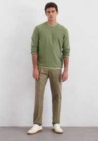 Jasnozielony sweter z prążkowanymi mankietami, w połączeniu z jasnymi khaki spodniami. Model stoi w białych sneakersach na neutralnym tle.