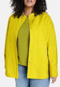 Žlutá bunda na knoflíky s kulatým výstřihem a náprsní kapsou, nošená přes zelený top, v kombinaci s modrými džíny. Hladká, lehká tkanina.