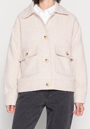 Beżowa teksturowana kurtka z dużym kołnierzem, cztery brązowe guziki, dwie patki na piersi, noszona na białej koszuli i czarnych spodniach.