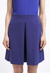 Fialová áčková sukně s pasem a záhyby, vyrobená z hladkého materiálu, která sahá nad kolena a má jednobarevný design.