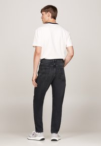 Jeans in denim nero con vestibilità dritta, cinque tasche e dettagli di cucitura sottili, abbinati a una maglietta bianca e sneakers grigie.