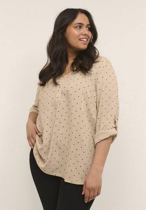 Chemise beige à pois noirs, tissu léger, décolleté en V, manches retroussées trois quarts et ourlet légèrement incurvé.