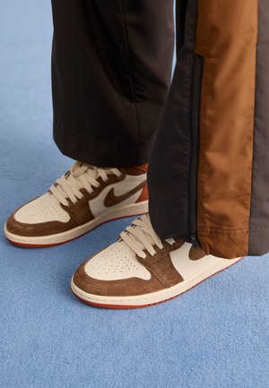 Sneakers Nike con una base in pelle bianca, sovrapposizioni in suede marrone, lacci beige e una suola in gomma rossa; abbinati a pantaloni sportivi marroni.