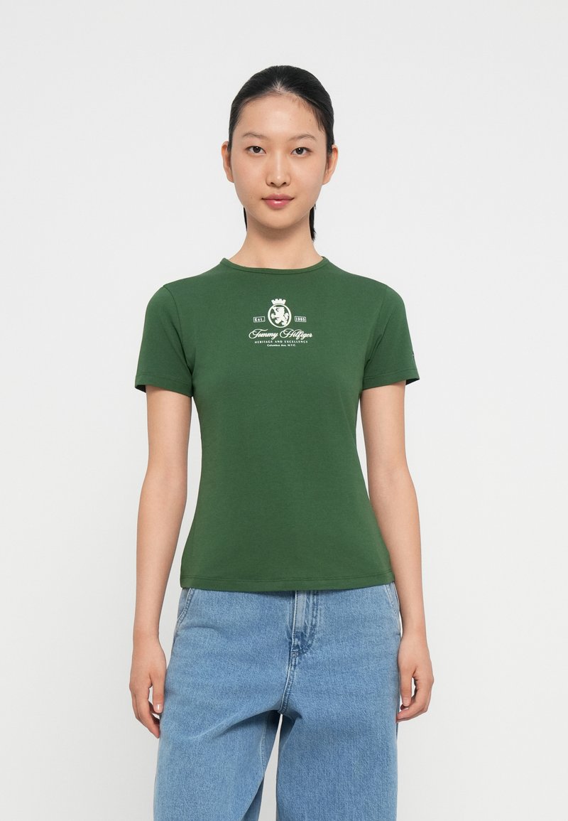 Camiseta de algodón verde con cuello redondo, mangas cortas y un estampado de logo blanco en el pecho, combinada con jeans de mezclilla azul claro.