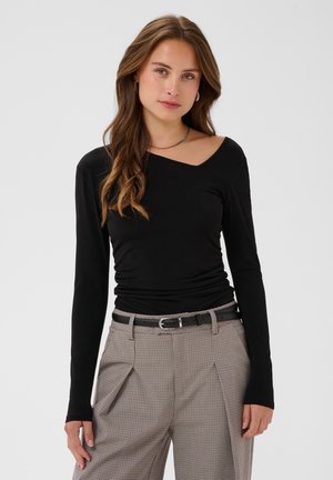 Femme aux longs cheveux bruns portant un haut noir asymétrique à manches longues et un pantalon à carreaux taille haute avec une ceinture noire, face à l'avant.