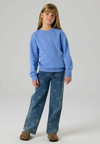 Mėlynas megztinis su gofruotomis rankogaliais ir kaklu, derinamas su plataus pjūvio džinsais su širdelių raštais ir subtilių dėvėjimosi žymių. Rudos spalvos avalynė.