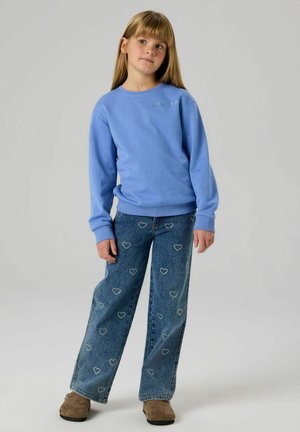 Blauer Pullover mit gerippten Bündchen und Rundhalsausschnitt, kombiniert mit weit geschnittenen Jeans mit Herzmustern und dezenten Used-Effekten. Beige Schuhe.