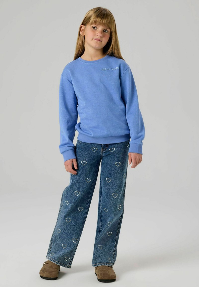 Mėlynas megztinis su gofruotomis rankogaliais ir kaklu, derinamas su plataus pjūvio džinsais su širdelių raštais ir subtilių dėvėjimosi žymių. Rudos spalvos avalynė.