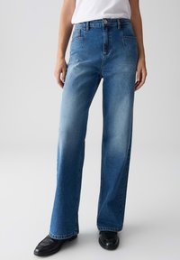 Donna che indossa jeans blu a vita alta, a gamba dritta, con aree sbiadite, mocassini neri e una maglietta bianca senza maniche infilata dentro su uno sfondo chiaro.