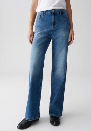 Femme portant un jean bleu taille haute, coupe droite avec des zones décolorées, des mocassins noirs et un top sans manches blanc rentré sur un fond clair.