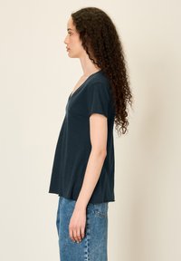 Temno modra majica z V-izrezom, kratkimi rokavi, sproščenega kroja in mehke teksture, v kombinaciji s svetlo modrimi hlačami, prikazano v bočni profilni perspektivi.