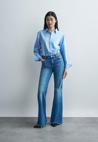 Kvinde står op ad en hvid væg iført en lyseblå skjorte med knapper, vide blå jeans med frynsede kanter og sorte spidse sko.