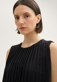 Čierny bezrukávový top s vertikálnymi skladmi, okrúhlym výstrihom a textúrovanou látkou. Obrázok sa sústreďuje na hornú časť tela a tvár modelky.