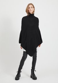 Poncho preto de malha com gola alta, apresentando uma forma triangular e textura macia, combinado com leggings de couro pretas e botas com atacadores.