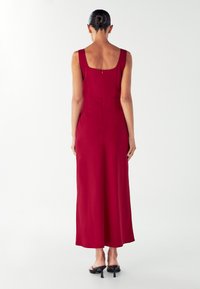 Dlhé, bezrukávové červené šaty s hranatým výstrihom na chrbte a priliehavým pásom. Hladká látka s dlhým lemom až po zem, doplnené čiernymi sandálmi na podpätku.