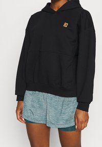 Schwarzer Pullover-Hoodie mit einer Vordertasche, strukturiertem Stoff und einem kleinen, mehrfarbigen Logo auf der Brust. Kombiniert mit melierten Shorts.