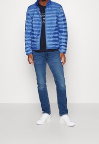 Blaue Steppjacke mit horizontalen Nähten, Reißverschluss und Stehkragen, getragen über einem dunklen Hemd und kombiniert mit blauen Jeans und weißen Sneakern.