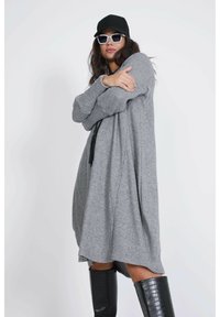 Grijze gebreide jurk met een losse pasvorm, lange mouwen en kwastaccenten. G gedragen met zwarte kniehoge laarzen met krokodillenprint en een pet.