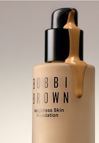 Bobbi Brown Weightless Skin Foundation i en frostet beige flaske med pumpe i sort. Væske drypper fra dispenseren og viser en kremet konsistens.