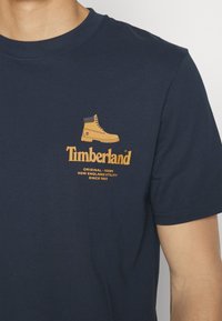 Tmavě modré bavlněné tričko s grafikou žluté boty Timberland a logem značky v oranžové barvě, s krátkými rukávy a kulatým výstřihem.