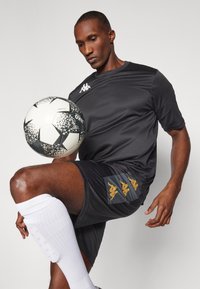 Sort træningsskjorte og shorts med gyldne accenter, parret med hvide sokker. En mønstret fodbold er balanceret på knæet.