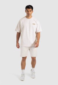 Conjunto de camiseta y pantalones cortos de algodón beige claro. Mangas cortas, ajuste holgado, logo bordado en el pecho. Combinado con zapatillas blancas y calcetines a rayas.