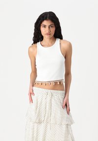 White ribbed crop top with a rounded neckline, featuring a decorative trim of shells and beads along the hem. Paired with a polka dot skirt.