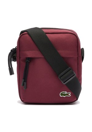 Bolso cruzado color marrón con una correa ajustable negra, cierre con cremallera y un pequeño logo verde de cocodrilo en la parte inferior frontal.