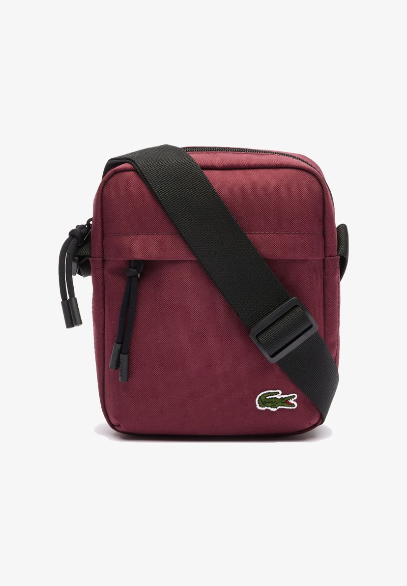 Bolso cruzado color marrón con una correa ajustable negra, cierre con cremallera y un pequeño logo verde de cocodrilo en la parte inferior frontal.