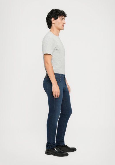 Muškarac s kovrčavom kosom stoji u profilu noseći sivi majicu, tamnoplave uske traperice i crne kožne cipele na bijeloj pozadini.