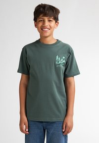 Groene katoenen T-shirt met korte mouwen, voorzien van een klein gedrukt ontwerp in lichtblauw op de linkerborst, gecombineerd met blauwe jeans.