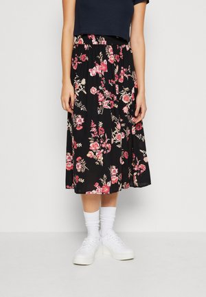 Femme portant une jupe midi noire avec imprimé floral rose, chaussettes blanches, baskets blanches et un top court noir, debout devant un fond uni.