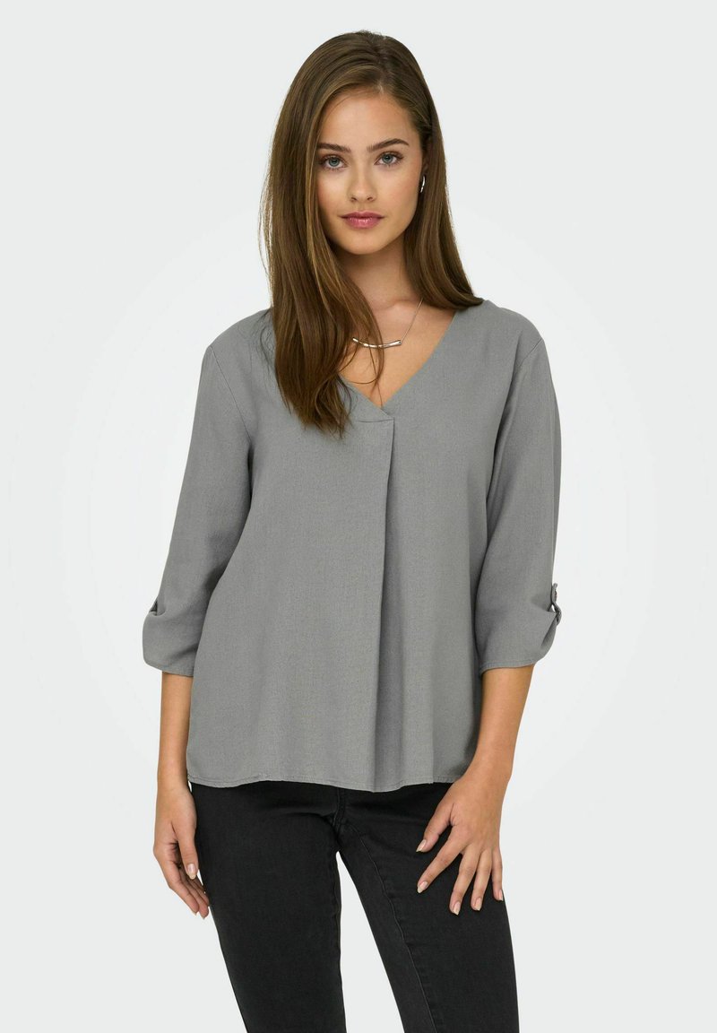 Blusa gris de cuello en V con mangas de tres cuartos, que presenta una textura suave y un corte holgado. Los detalles notables incluyen puños enrollados y un diseño plisado.