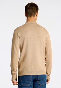 Beige långärmad tröja med krage, slät textur, ribbade ärmslut och fåll. Bakifrån visas en figursydd design med sidslitsar.