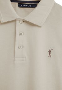 Polo beige avec trois boutons, un col classique, un petit logo de golfeur brodé sur la poitrine et une étiquette "Polo Club" à l'intérieur du col.