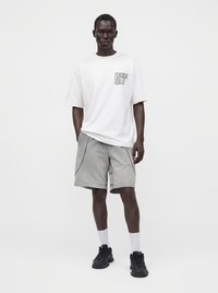Homme portant un t-shirt blanc oversize avec le texte "OFF", un short gris avec passepoil noir, des chaussettes blanches et des baskets noires, debout devant un fond uni.