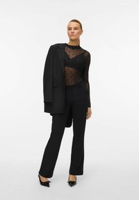 Zwarte pak met een op maat gemaakt colbert, doorzichtige top met stippen, strakke broek en puntige hakken. Soepele stof met een gestructureerd ontwerp.