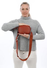 Hall üleval kasukas halli kõrge kaelusega kampsuniga, hoides väike pruuni nahast õlakotti reguleeritava rihmaga, valge taustaga.