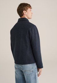 Jachetă din lân�ă bleumarin cu guler rabatabil, țesătură texturată și croi drept. Asortată cu blugi din denim de un albastru deschis, văzută din spate.