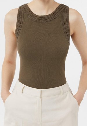 Haut sans manches en maille vert olive avec bordure texturée, porté rentré dans un pantalon blanc taille haute, mains reposant dans les poches.