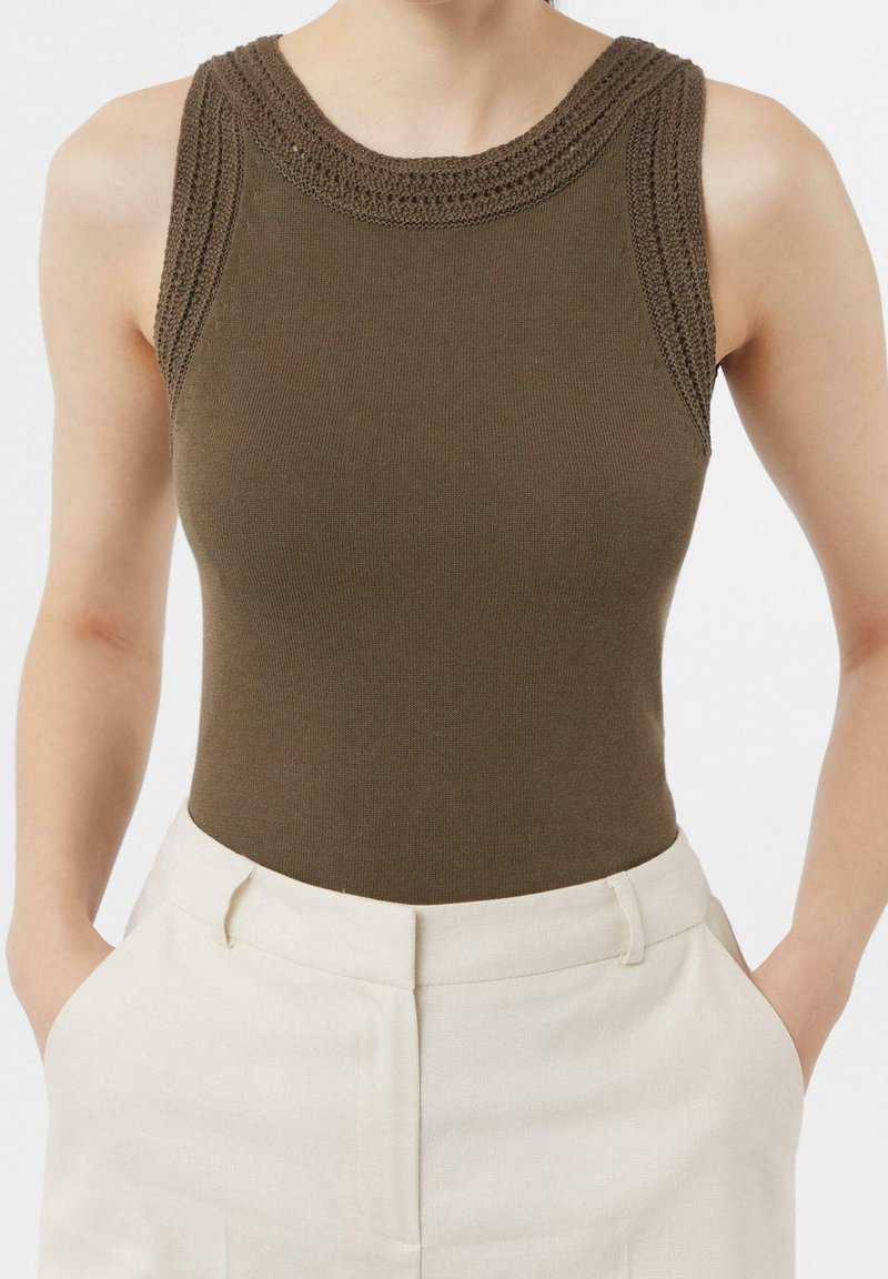 Haut sans manches en maille vert olive avec bordure texturée, porté rentré dans un pantalon blanc taille haute, mains reposant dans les poches.