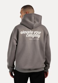 Sudadera con capucha gris que presenta una gran impresión blanca con la frase "elevate your everyday" en la parte posterior; fabricada en una tela suave, de corte relajado.
