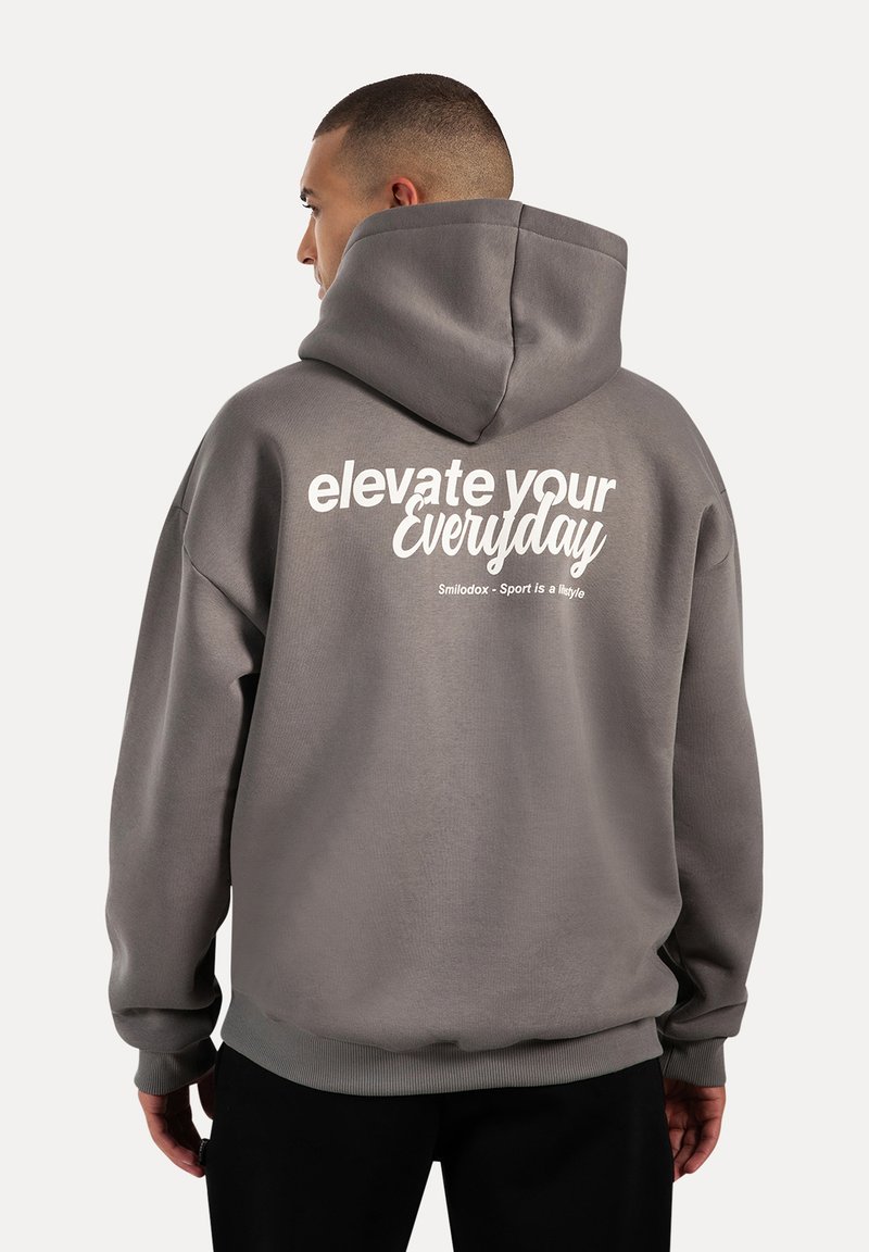Sudadera con capucha gris que presenta una gran impresión blanca con la frase "elevate your everyday" en la parte posterior; fabricada en una tela suave, de corte relajado.