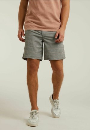 Homme portant un short à carreaux gris, une chemise à manches courtes rose clair et des baskets blanches, sur fond uni clair.