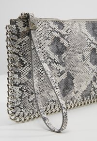 Pochette avec motif en peau de serpent gris et noir, bordure en chaîne argentée et bracelet assorti posée sur une surface blanche.