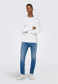 Bijela košulja dugih rukava, plave traperice i bijeli tenisice. Košulja je jednostavna, traperice su malo izblijedjele, a tenisice imaju suptilne detalje.