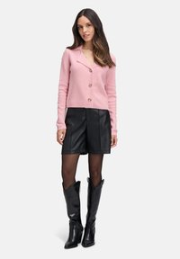 Frau in einer rosa, knöpfbaren Strickjacke, schwarzen Leder-Shorts, durchsichtigen schwarzen Strumpfhosen und schwarzen knielangen Stiefeln, die vor einem weißen Hintergrund steht.