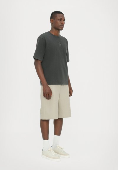Kortærmet, mørkegrå bomulds T-shirt med et lille logo, parret med beige shorts. Båret med hvide sokker og lyse sneakers.