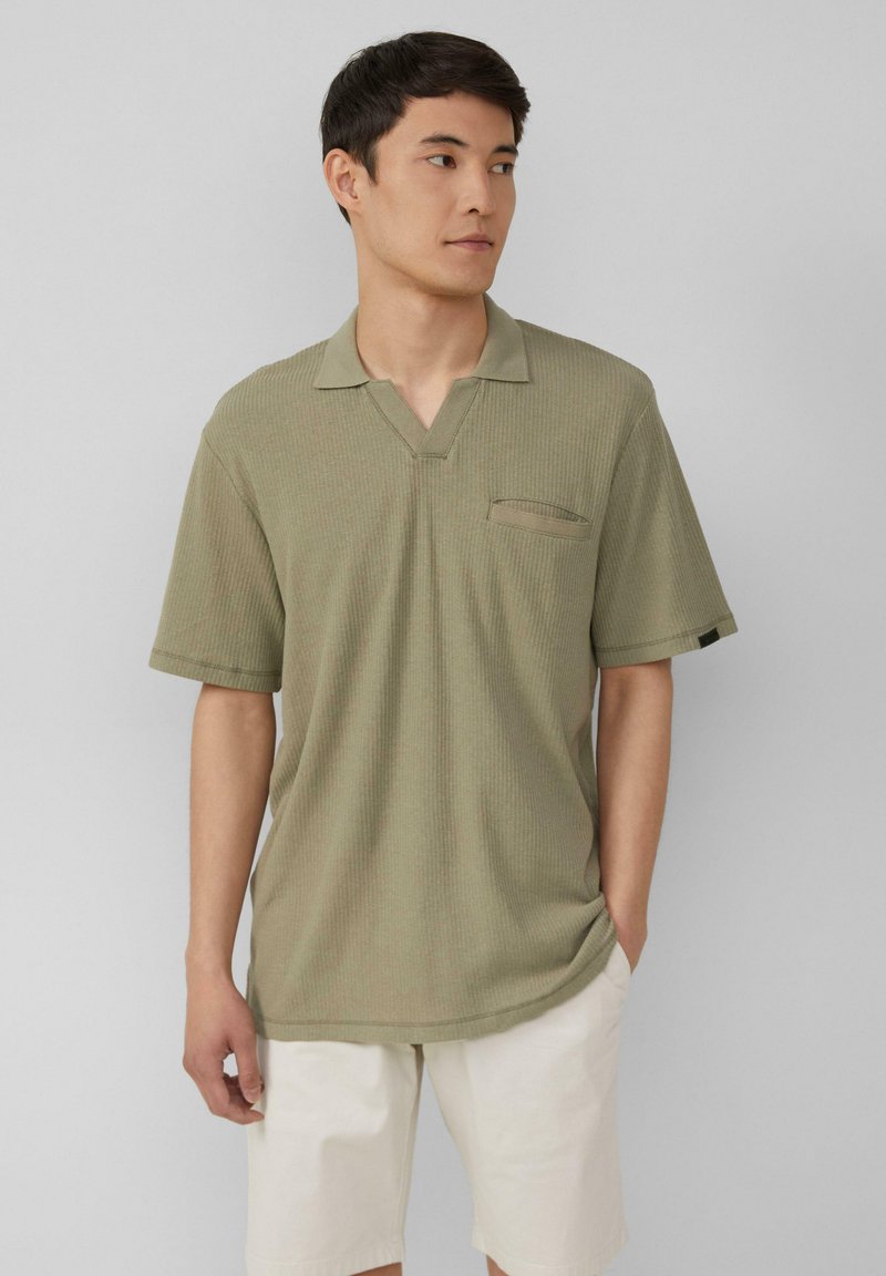 Oliwkowa koszulka polo z krótkim rękawem w ryflowanym wzorze, z dekoltem w kształcie litery V oraz kieszonką na piersi, w połączeniu z jasnymi szortami.